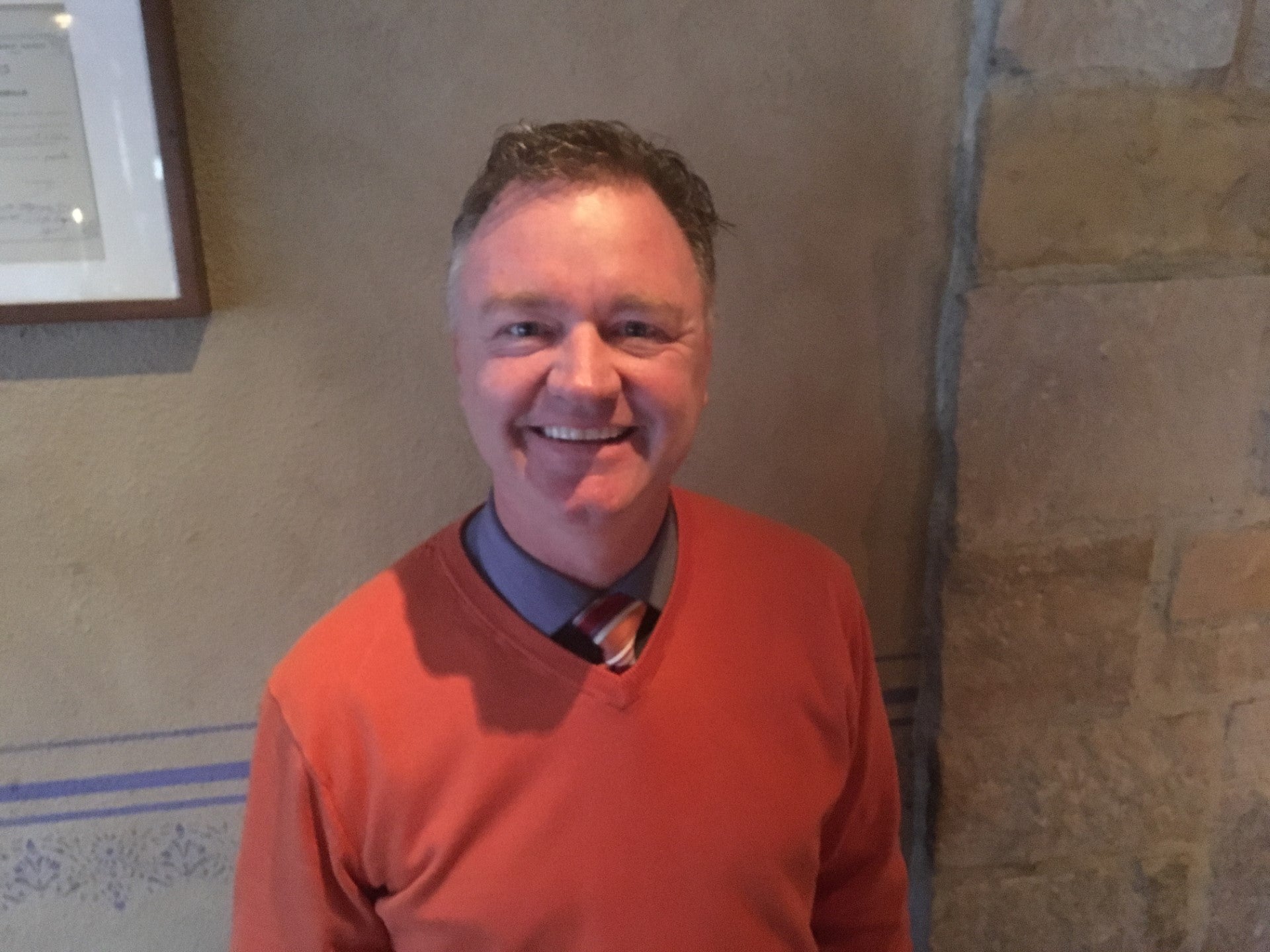 Man wearing an orange sweater with a blue shirt underneath, standing against a stone wall.